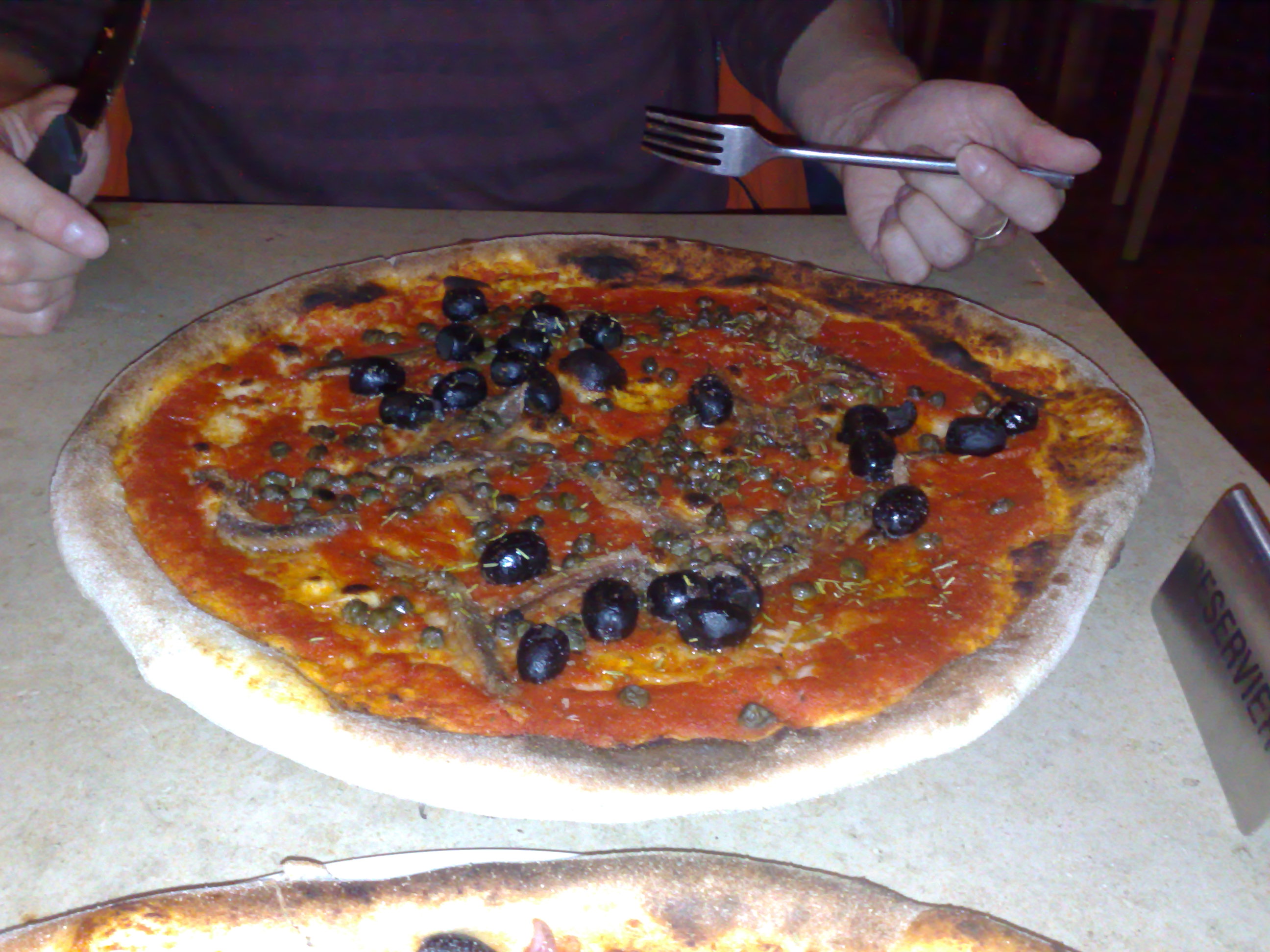 Unter dieser Pizza befindet sich ein großer Teller. Es ist eine Siciliana ohne Käse ...aber mit Kapern, Sardellen und Oliven.