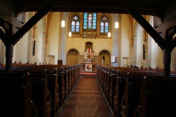 Unter der Empore angekommen öffnet sich der Blick auf den Kirchenraum, den Altar und den flachen Chorabschluss mit dem großen Drillingsfenster, dessen Originalverglasung aus der Bauzeit vom September 1899 bis Oktober 1900 erhalten geblieben ist. Auch die Kirchenbänke stammen noch aus dieser Zeit. Das Lilienmotiv aus den Terrakotta- Fußbodenfliesen wiederholt sich mehrfach in der Kirche.